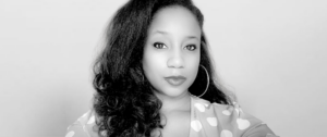 Portrait of a woman with long, wavy hair wearing large hoop earrings, facing the camera in a heart-patterned top (black and white).
