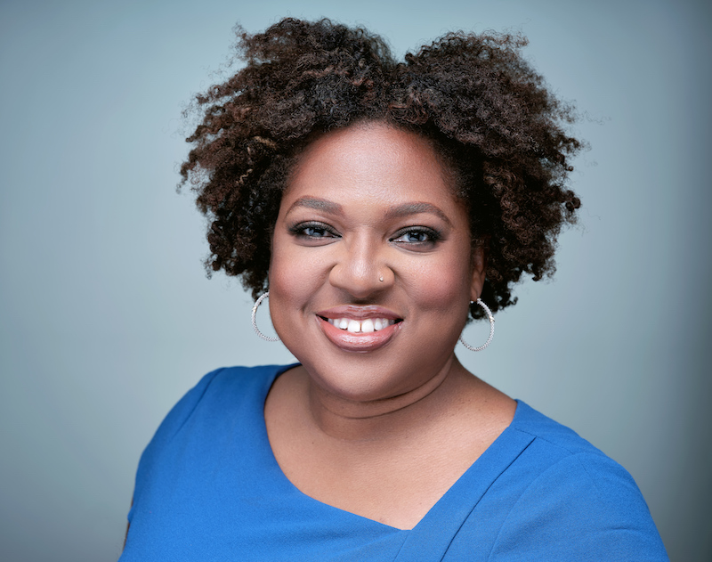 Smiling woman with curly hair in a blue top, posing for a professional headshot against a soft blue background.